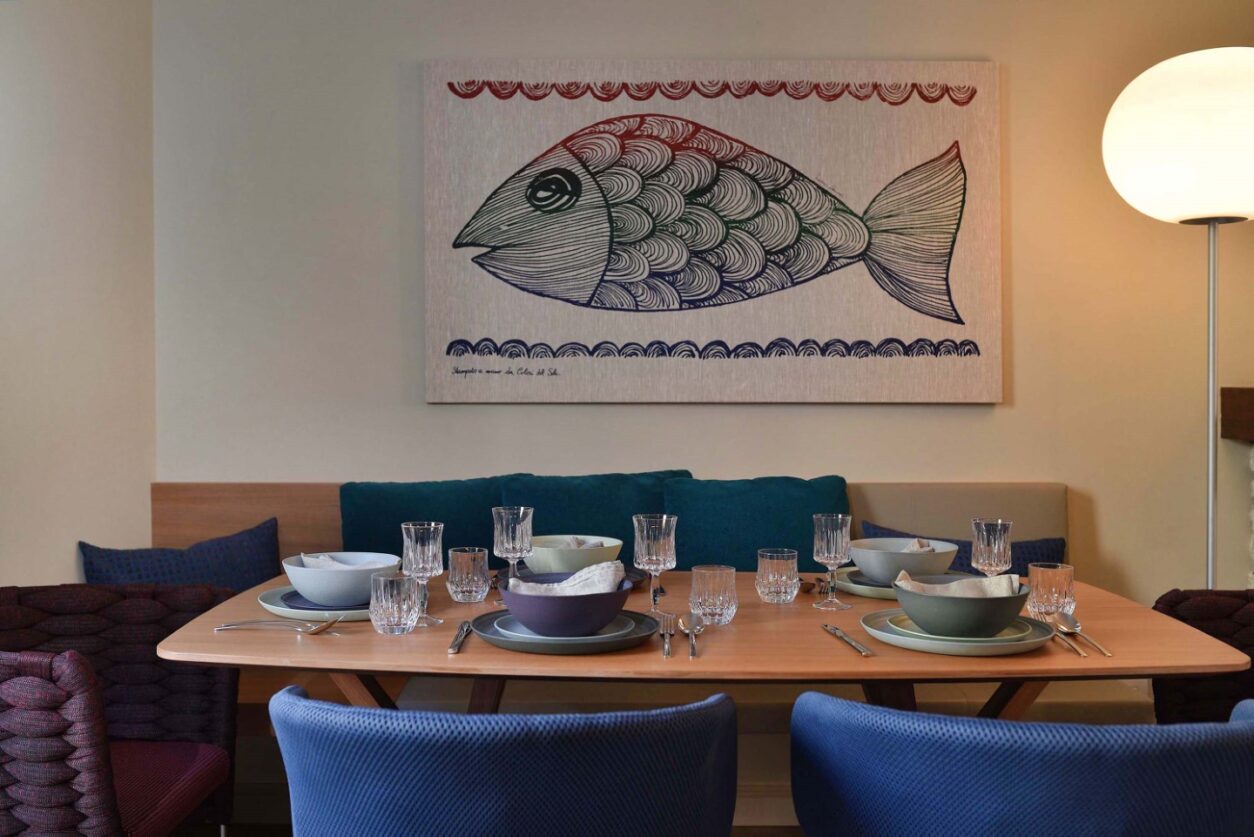 Im Refugium Serenitas steht der Esstisch mit blauen Stühlen und Sitzbank vor einem Wandbild mit maritimen Motiven