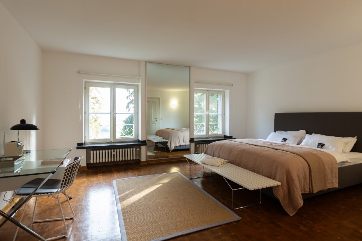 Ein großes Schlafzimmer in Brauntönen mit Doppelbett, Parkettboden und zwei Fenstern zum Garten im Haus Fürstenberg